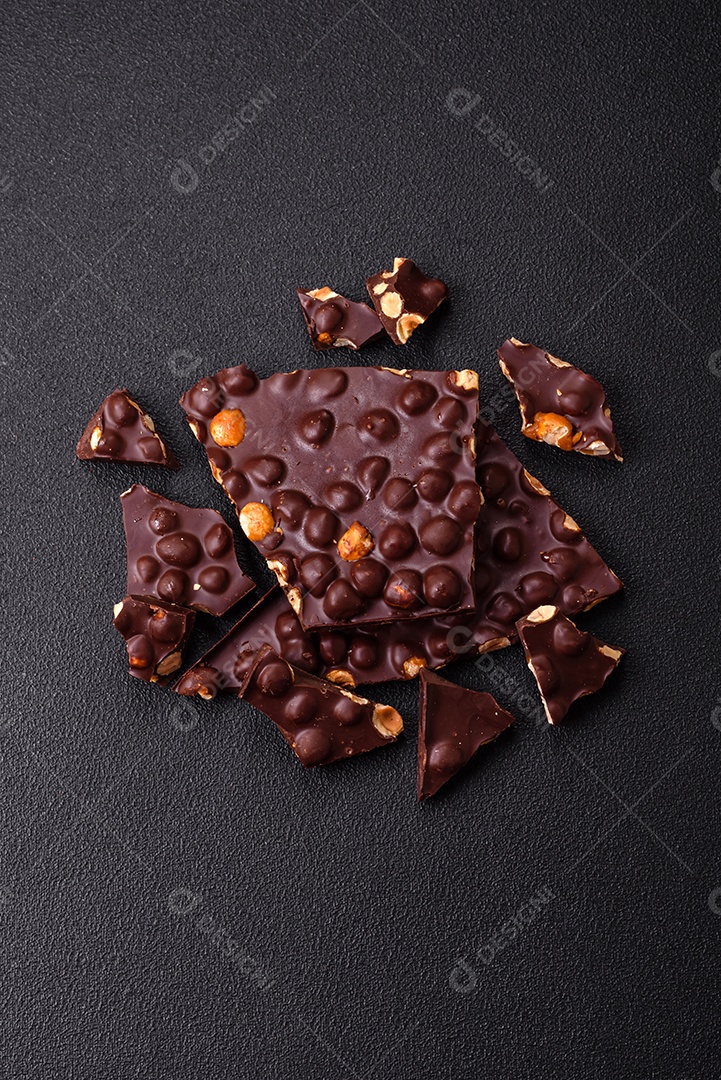 Chocolate amargo preto com avelãs inteiras, frutas cristalizadas e hortelã, quebrado em pedaços sobre fundo escuro de concreto