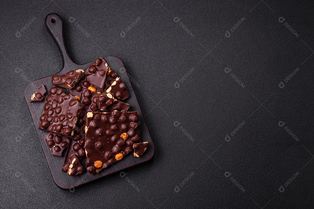 Chocolate amargo preto com avelãs inteiras, frutas cristalizadas e hortelã, quebrado em pedaços sobre fundo escuro de concreto
