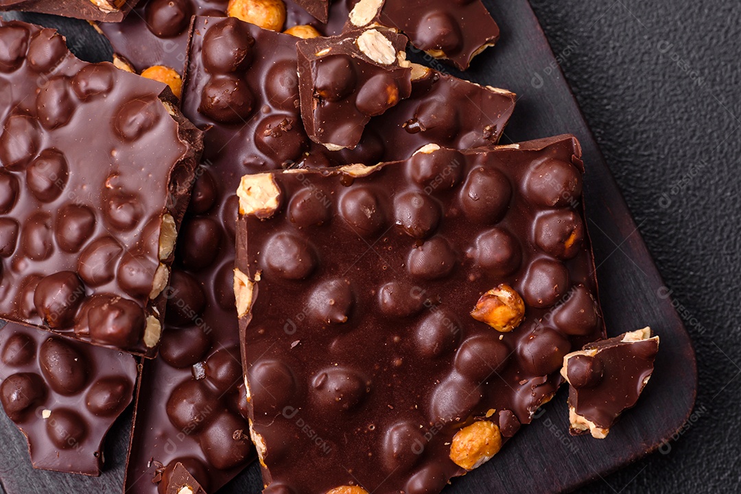 Chocolate amargo preto com avelãs inteiras, frutas cristalizadas e hortelã, quebrado em pedaços sobre fundo escuro de concreto