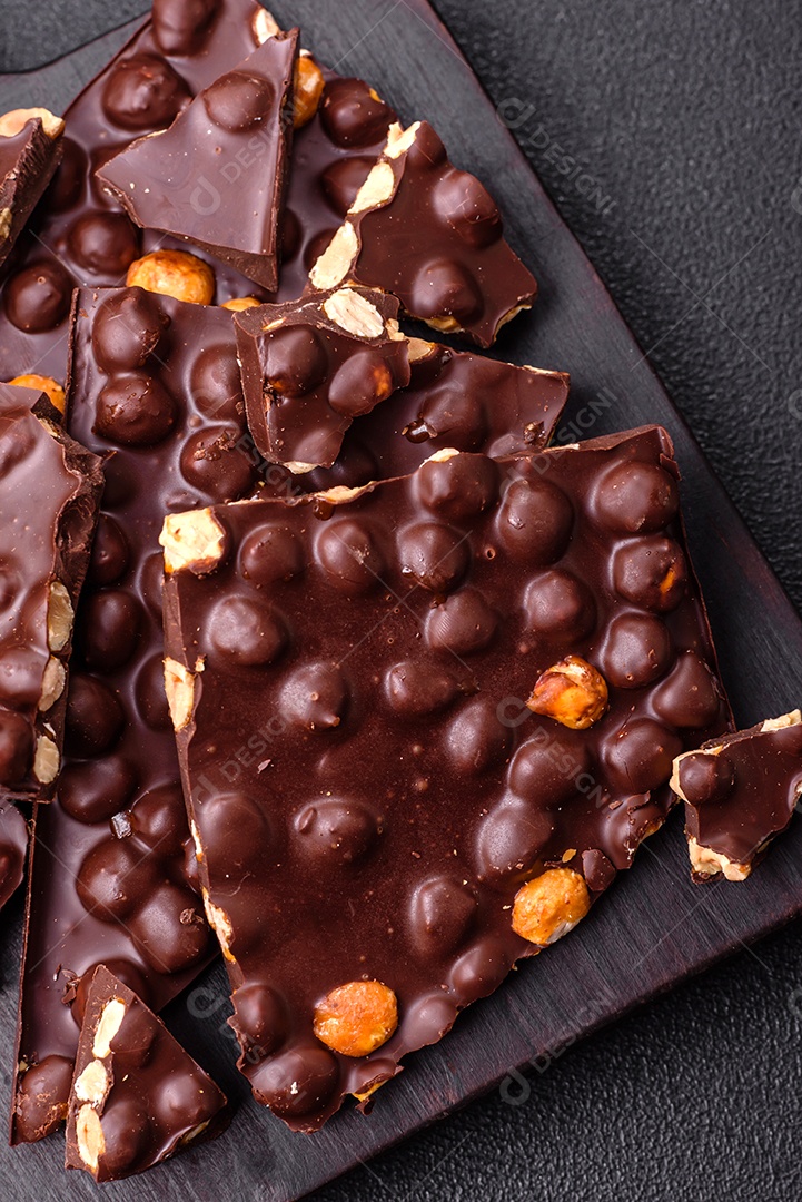 Chocolate amargo preto com avelãs inteiras, frutas cristalizadas e hortelã, quebrado em pedaços sobre fundo escuro de concreto
