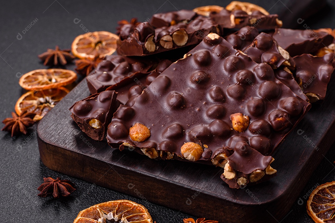 Chocolate amargo preto com avelãs inteiras, frutas cristalizadas e hortelã, quebrado em pedaços sobre fundo escuro de concreto