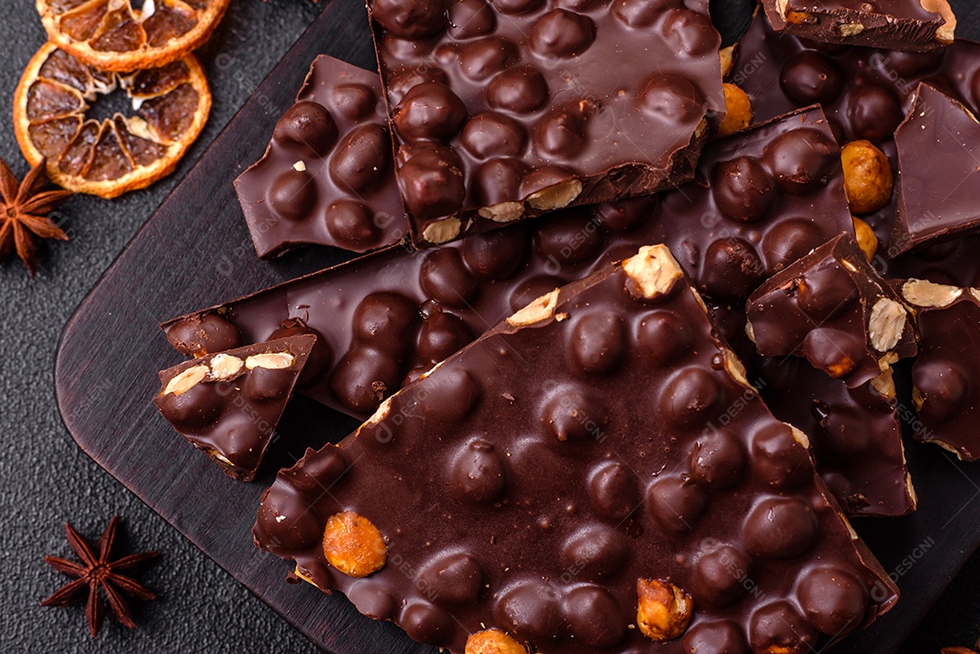 Chocolate amargo preto com avelãs inteiras, frutas cristalizadas e hortelã, quebrado em pedaços sobre fundo escuro de concreto