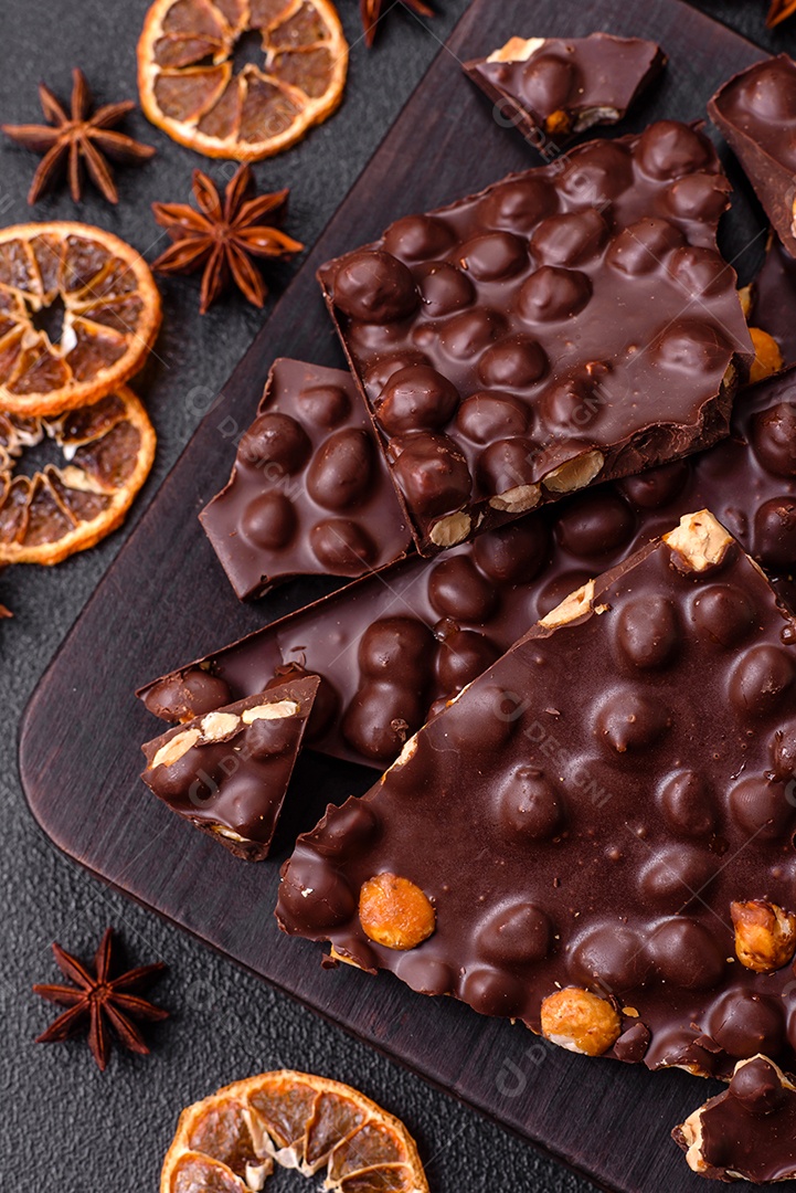Chocolate amargo preto com avelãs inteiras, frutas cristalizadas e hortelã, quebrado em pedaços sobre fundo escuro de concreto