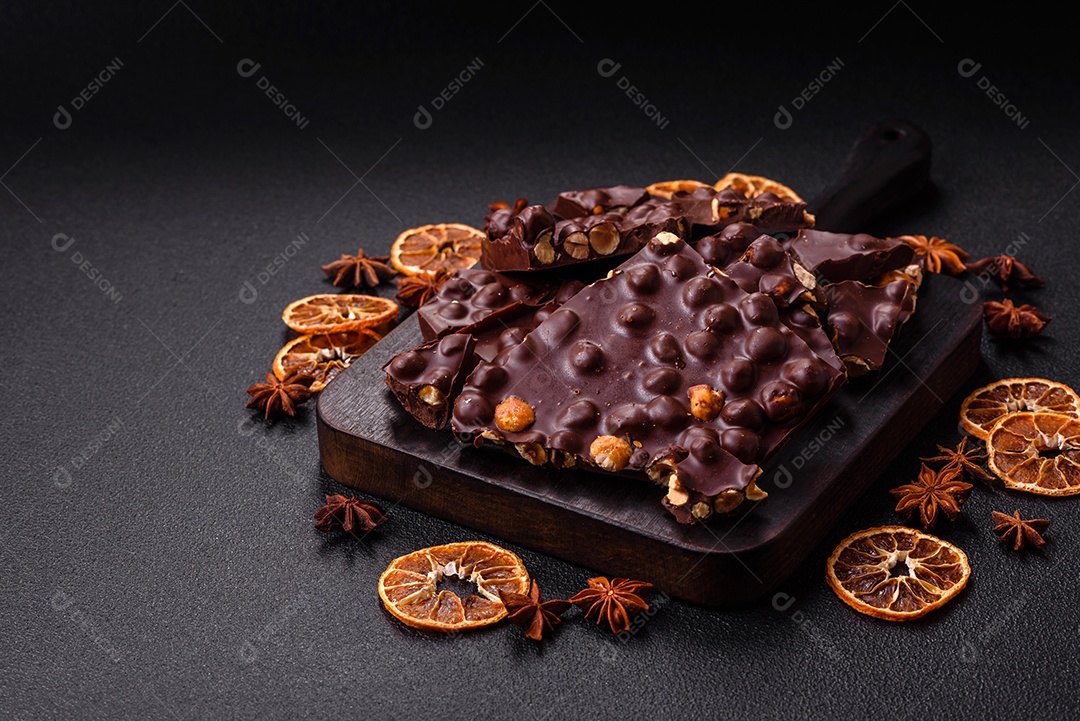 Chocolate amargo preto com avelãs inteiras, frutas cristalizadas e hortelã, quebrado em pedaços sobre fundo escuro de concreto