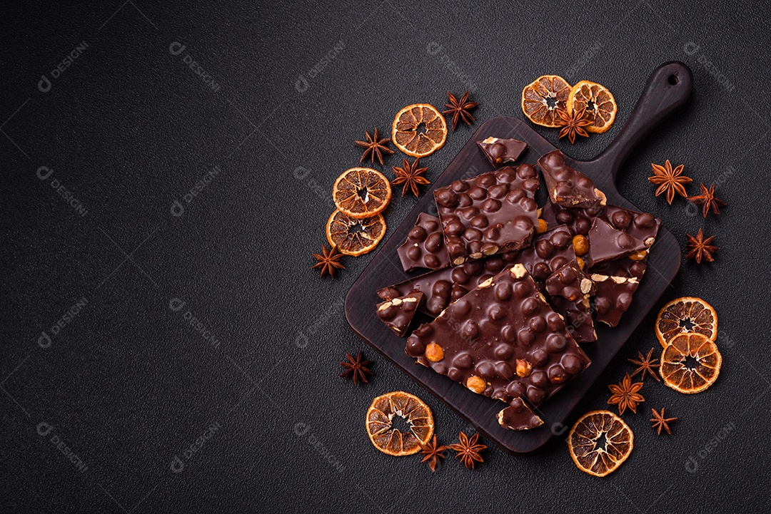 Chocolate amargo preto com avelãs inteiras, frutas cristalizadas e hortelã, quebrado em pedaços sobre fundo escuro de concreto