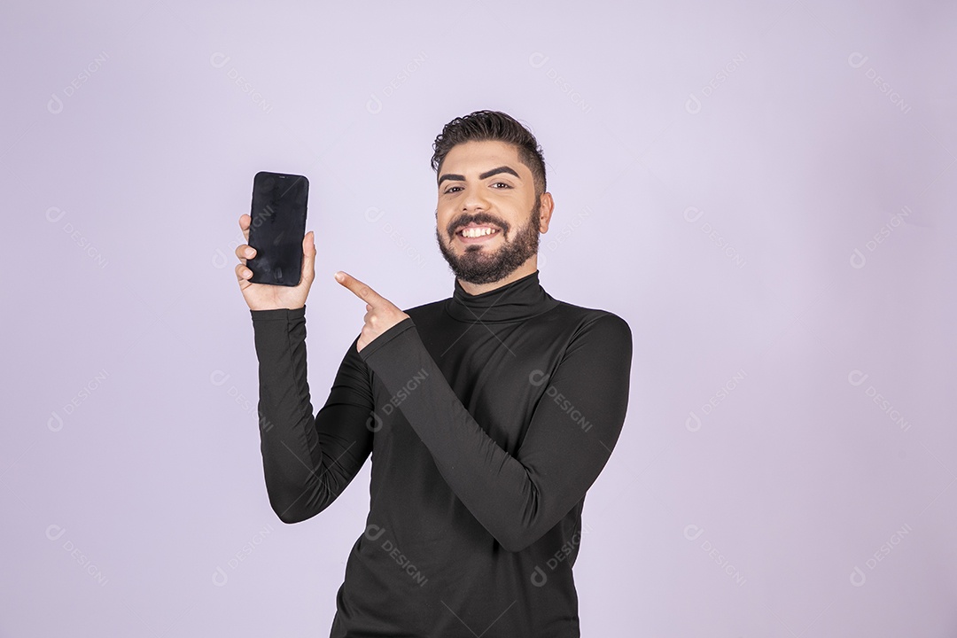 Homem sorrindo com celular nas mãos e gesto para o aparelho