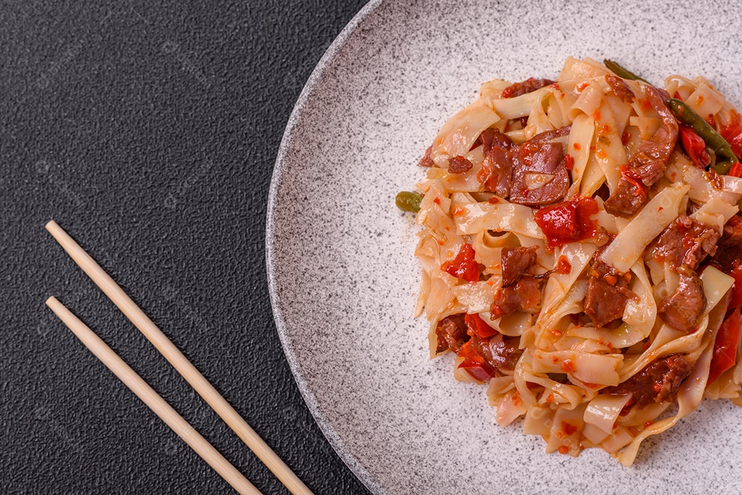 Delicioso prato de udon asiático com legumes, carne, sal e especiarias numa placa cerâmica sobre um fundo escuro de concreto