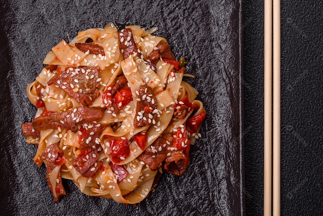 Delicioso prato de udon asiático com legumes, carne, sal e especiarias numa placa cerâmica sobre um fundo escuro de concreto