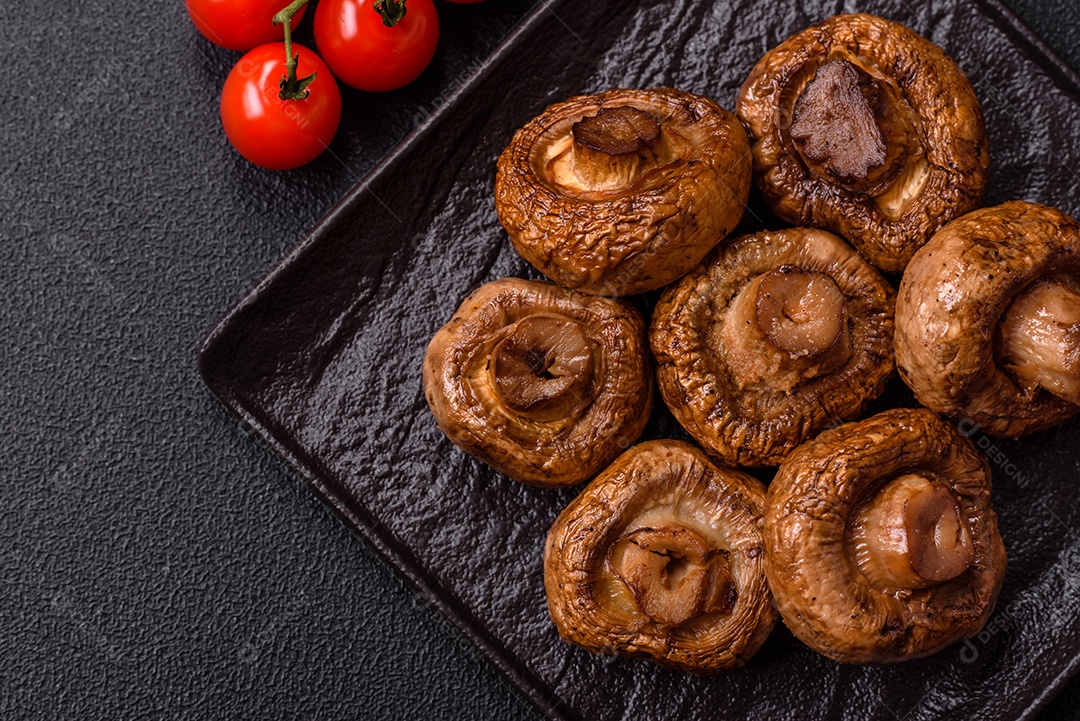 Deliciosos champignons inteiros e suculentos assados ​​na grelha com sal e especiarias em uma placa de cerâmica sobre um fundo escuro de concreto