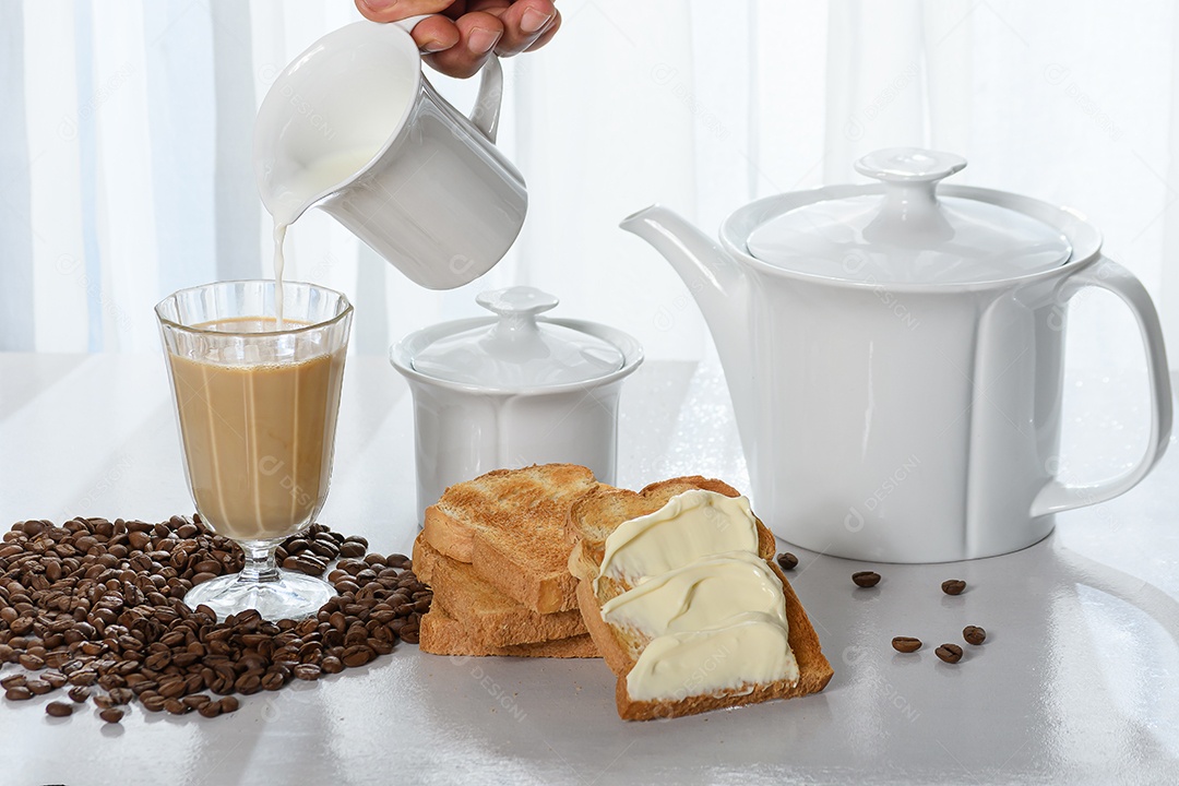 Mão colocando leite sobre uma xícara e torradas com requeijão ao lado