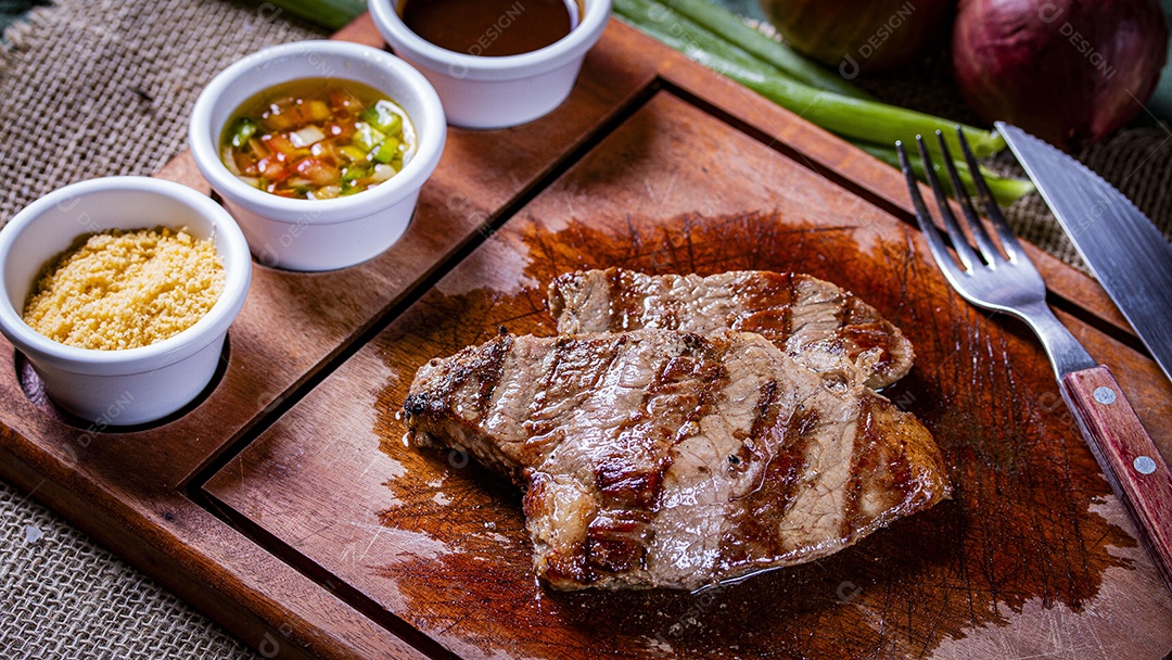 Carne assada sobre tábua de madeira com molho vinagrete farofa