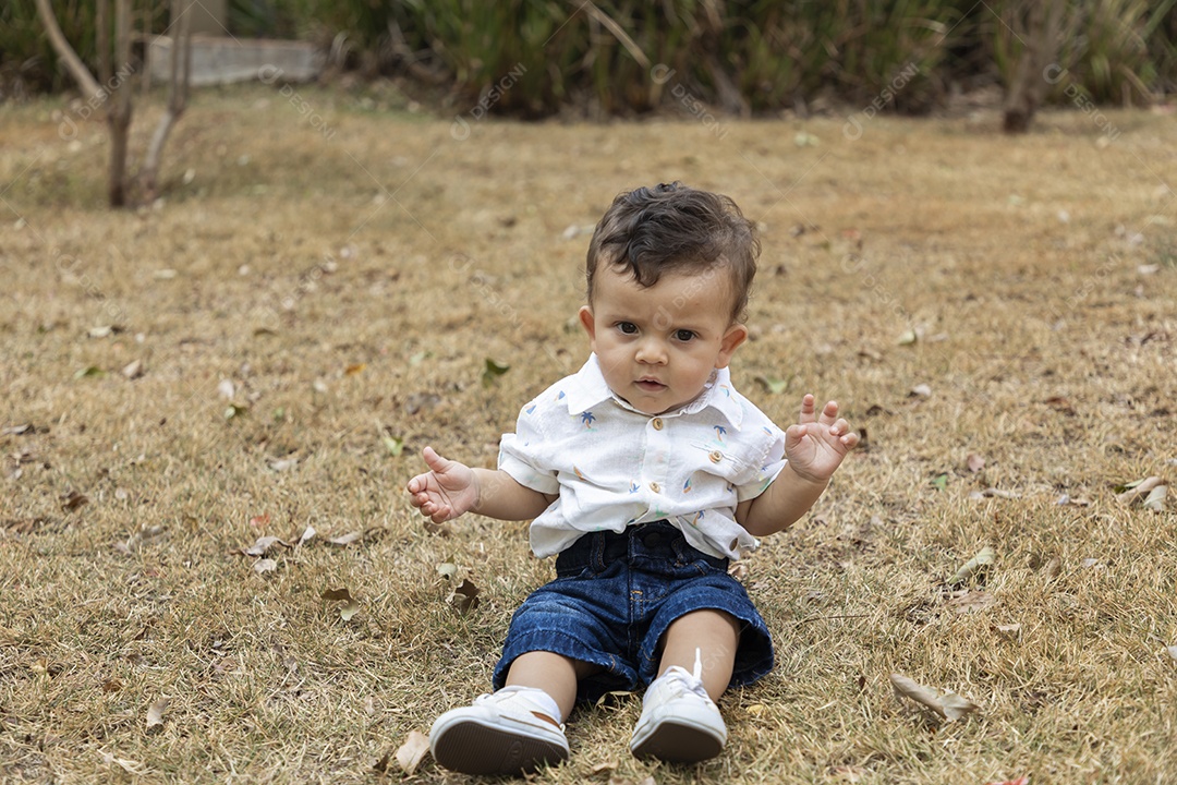Bebê de cara fechadinha sentado na grama de um parque