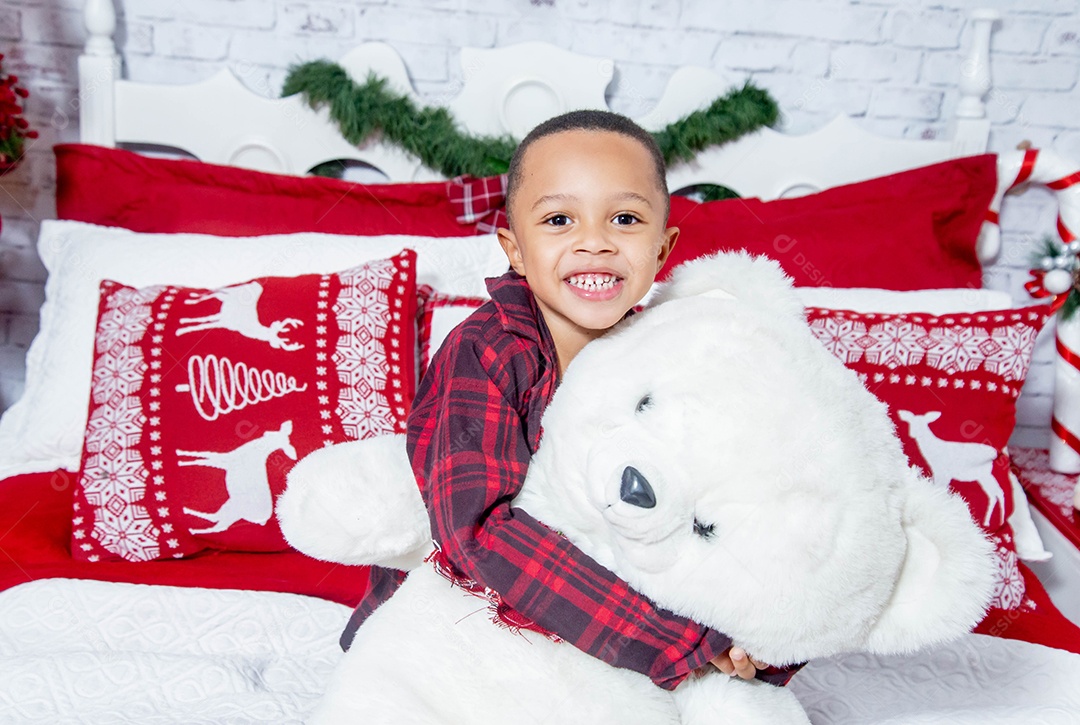 Garotinho lindo com urso e feliz numa cama decorada para o natal