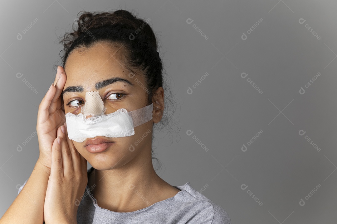 Mulher com mãos no rosto e nariz com curativo