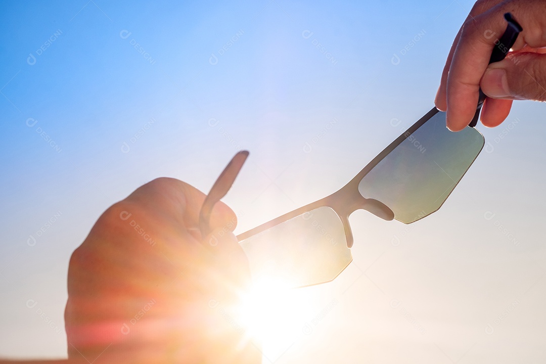 Close up Mão segurando óculos de sol esportivos elegantes contra o céu brilhante e o fundo do sol