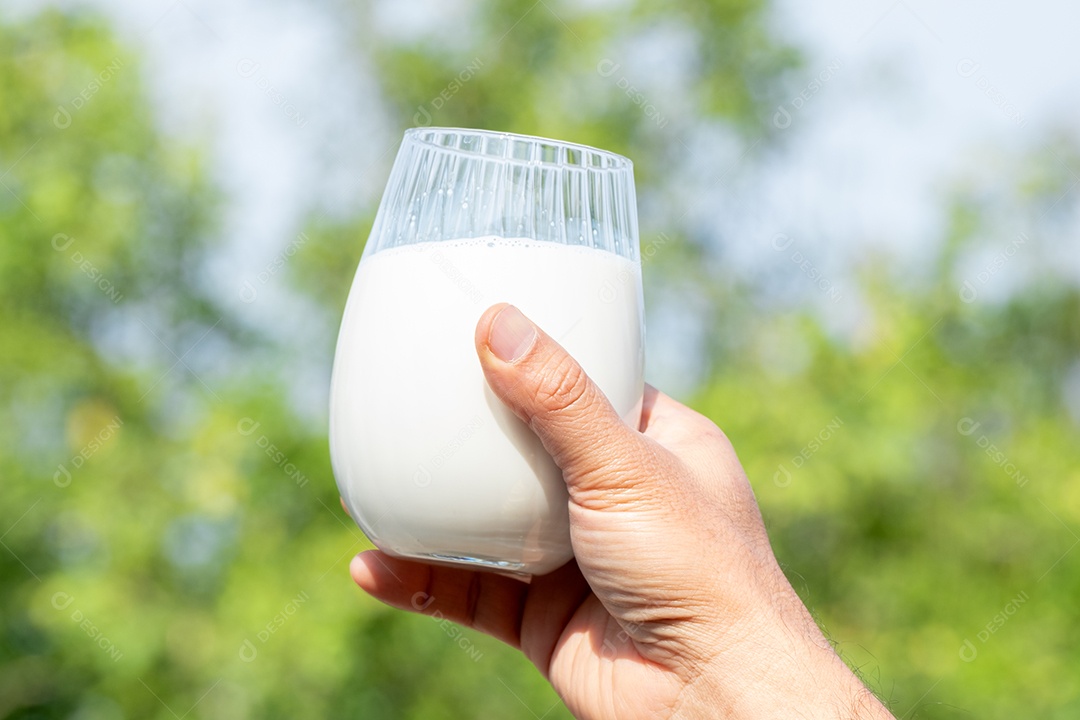 Mão de mãe segurando um copo de leite branco orgânico fresco com fundo desfocado verde da natureza e luz solar suave pela manhã