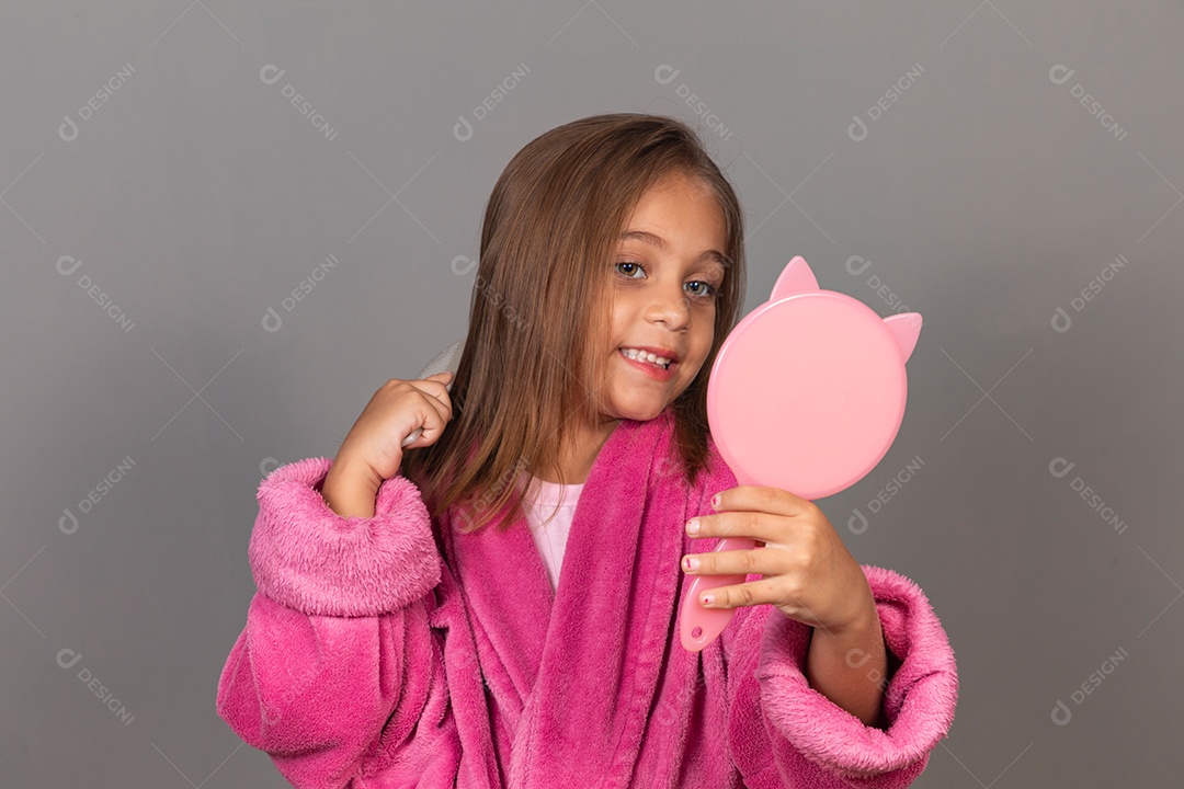 Linda menina usando roupão de banho sobre fundo rosa do isolado segurando espelho escovando o cabelo