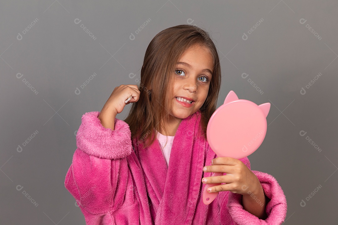 Linda menina usando roupão de banho sobre fundo rosa do isolado segurando espelho escovando o cabelo