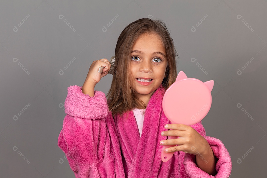Linda menina usando roupão de banho sobre fundo rosa do isolado segurando espelho escovando o cabelo