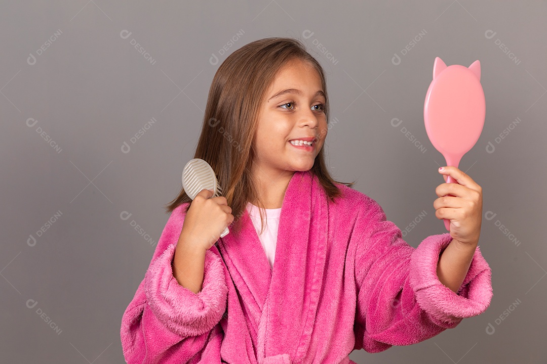 Linda menina usando roupão de banho sobre fundo rosa do isolado segurando espelho escovando o cabelo