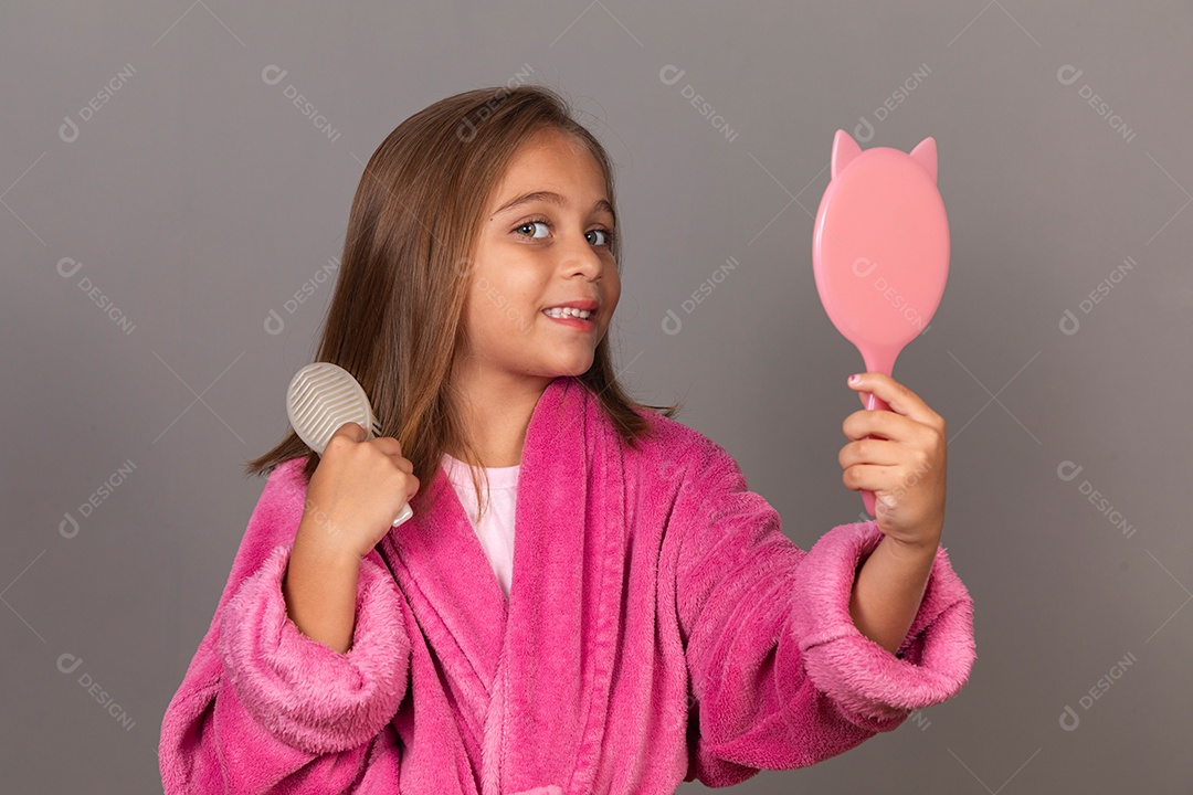 Linda menina usando roupão de banho sobre fundo rosa do isolado segurando espelho escovando o cabelo