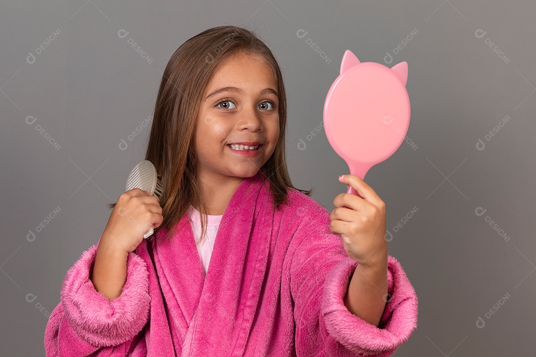 Linda menina usando roupão de banho sobre fundo rosa do isolado segurando espelho escovando o cabelo