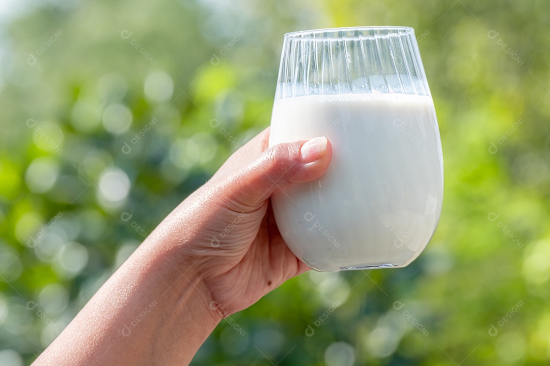 Mão de mãe segurando um copo de leite branco orgânico fresco com fundo desfocado verde da natureza e luz solar suave pela manhã