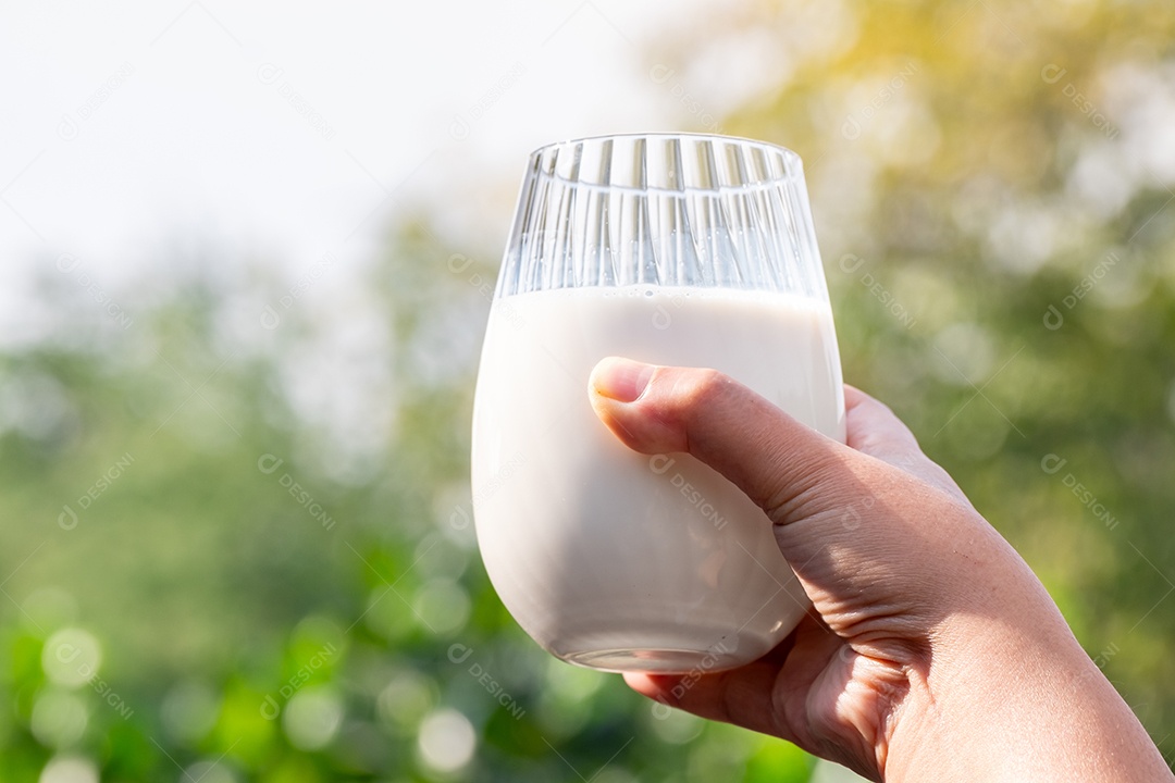 Mão de mãe segurando um copo de leite branco orgânico fresco com fundo desfocado verde da natureza e luz solar suave pela manhã