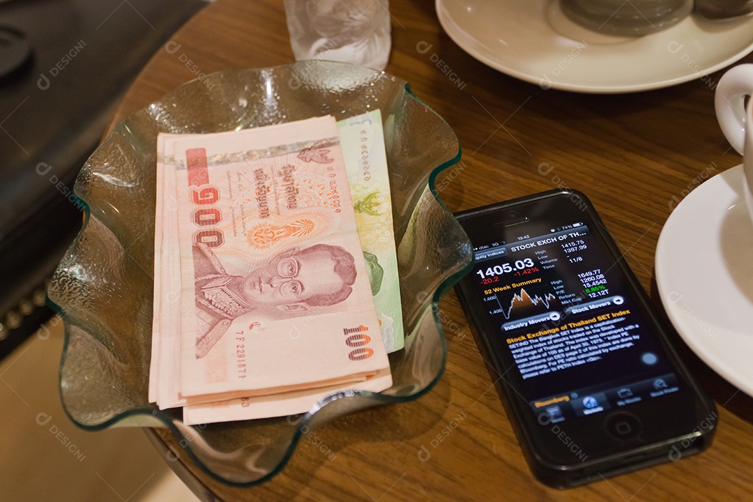 Nota de banco tailandesa em tijela ao lado de smrtphone com tecnologia avançada para investimento sobre mesa de madeira