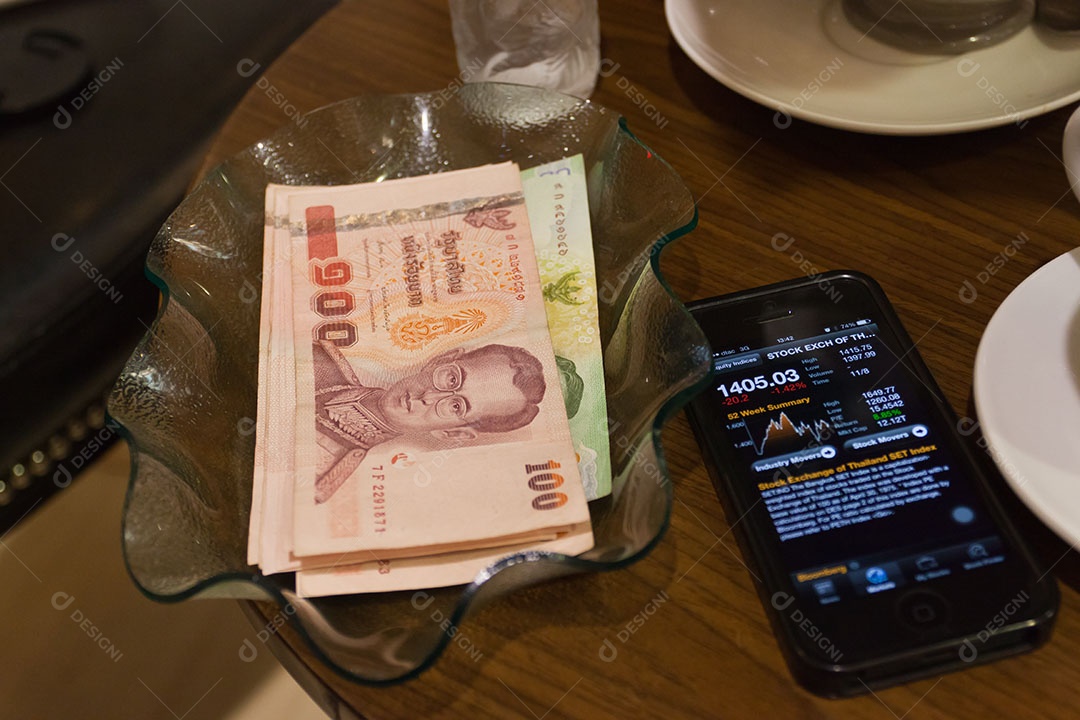 Nota de banco tailandesa em tijela ao lado de smrtphone com tecnologia avançada para investimento sobre mesa de madeira