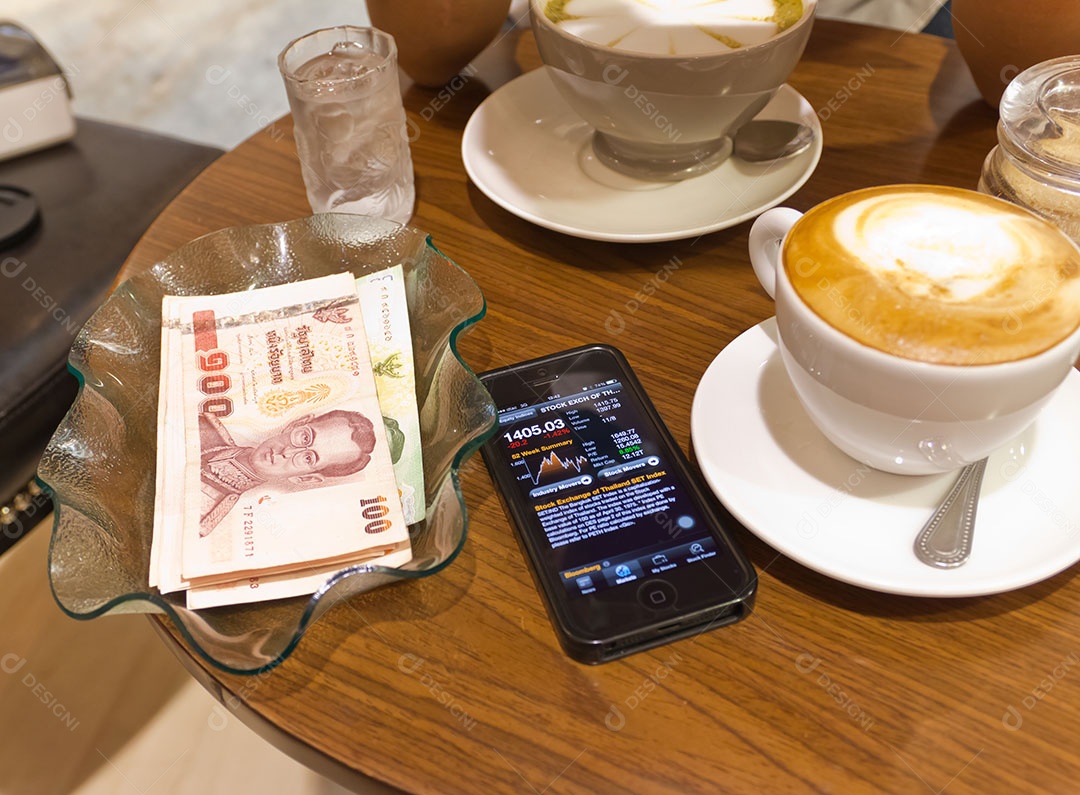 Nota de banco tailandesa em tijela ao lado de smrtphone com tecnologia avançada para investimento e xícara de cappuccino sobre mesa de madeira