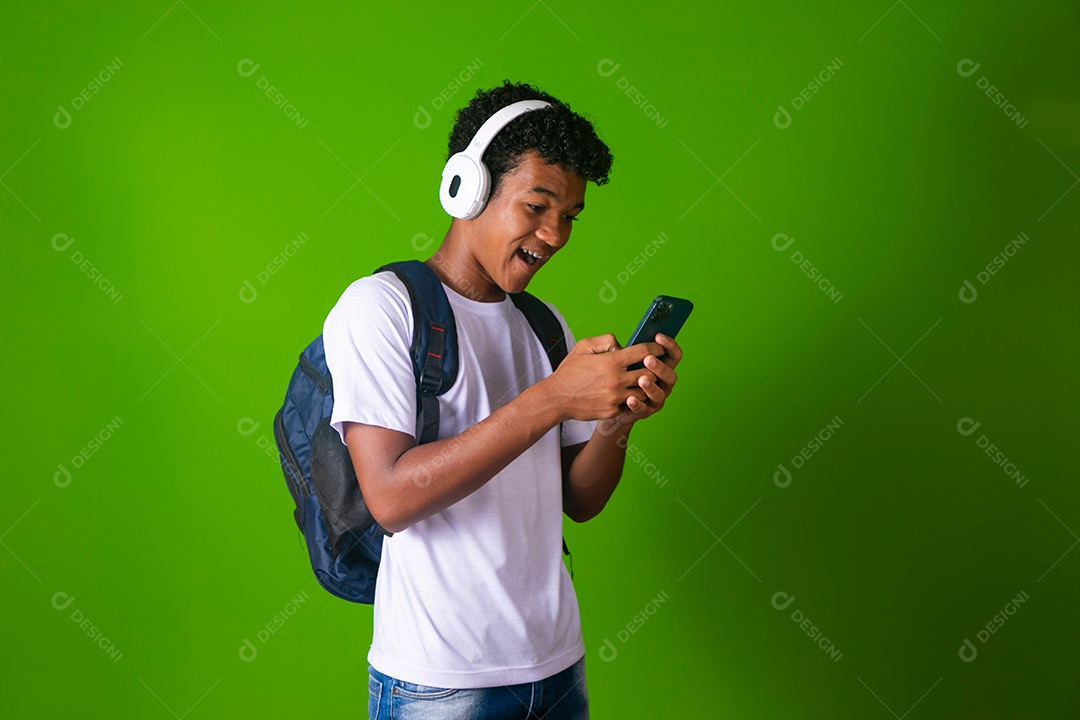 Lindo garoto negro usando fone de ouvido e mochila e segurando smartfhone sobre fundo verde isolado