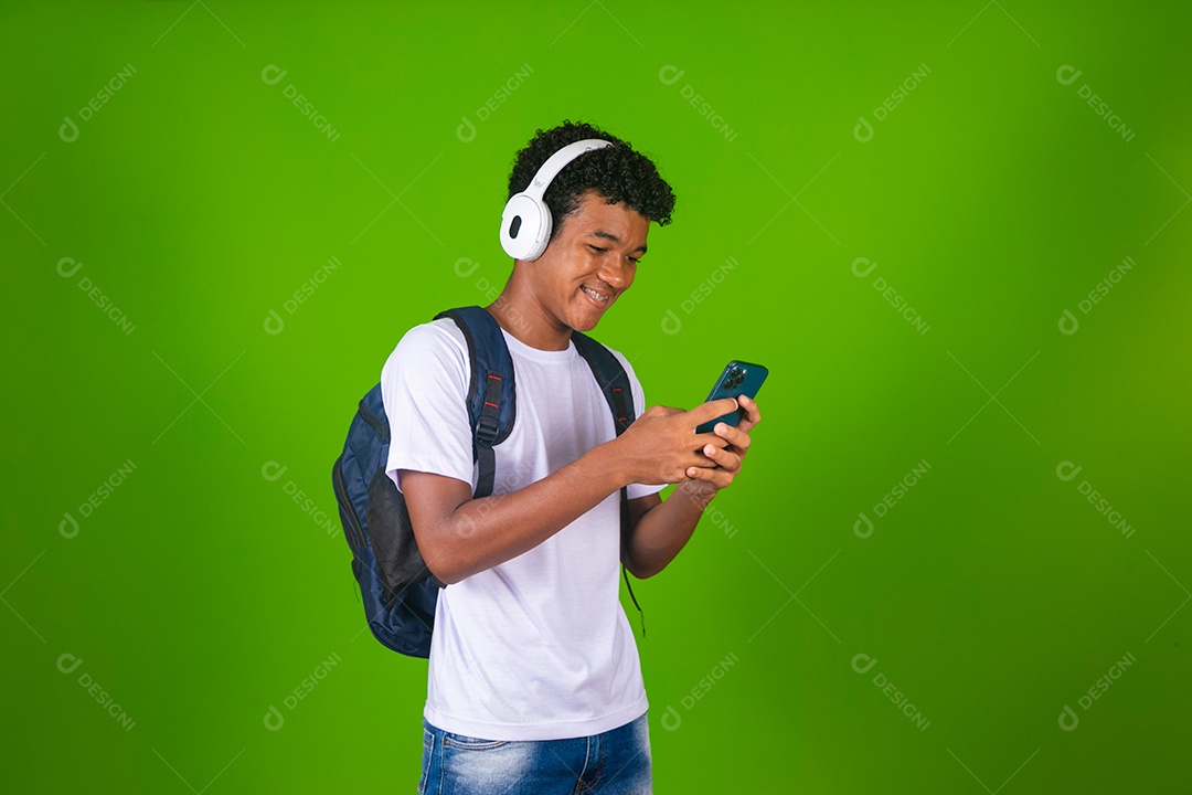 Lindo garoto negro usando fone de ouvido e mochila e segurando smartfhone sobre fundo verde isolado