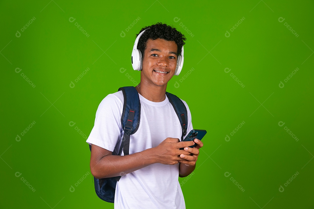 Lindo garoto negro usando fone de ouvido e mochila e segurando smartfhone sobre fundo verde isolado
