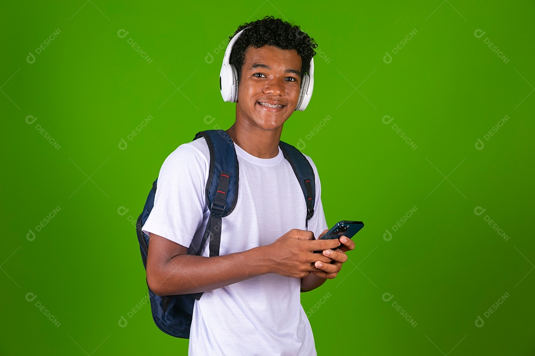 Lindo garoto negro usando fone de ouvido e mochila e segurando smartfhone sobre fundo verde isolado