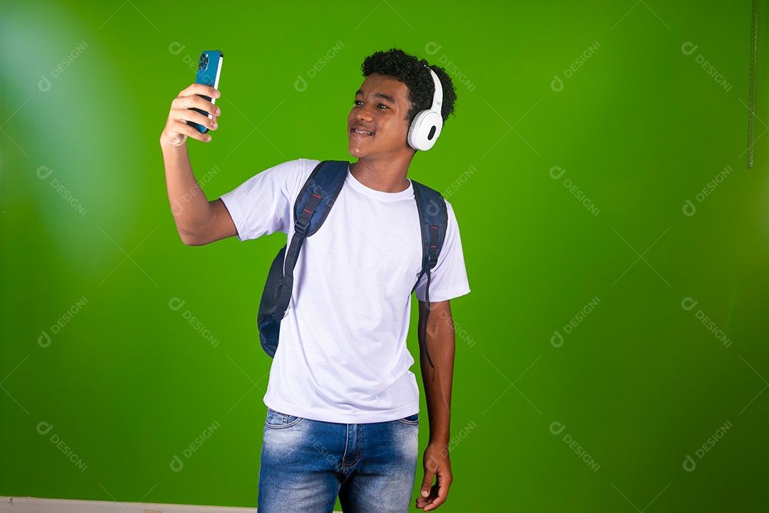 Lindo garoto negro usando fone de ouvido e mochila e segurando smartfhone sobre fundo verde isolado