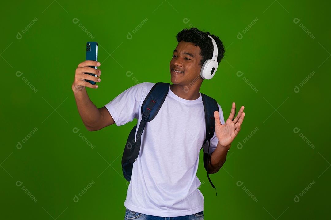 Lindo garoto negro usando fone de ouvido e mochila e segurando smartfhone sobre fundo verde isolado