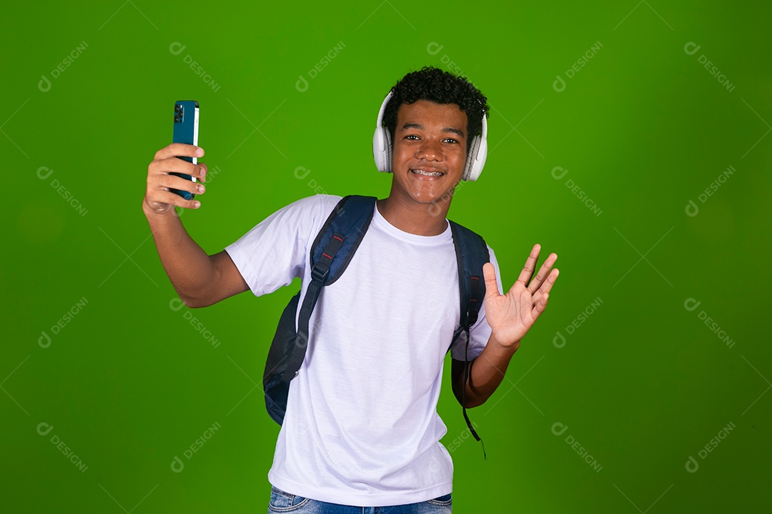 Lindo garoto negro usando fone de ouvido e mochila e segurando smartfhone sobre fundo verde isolado