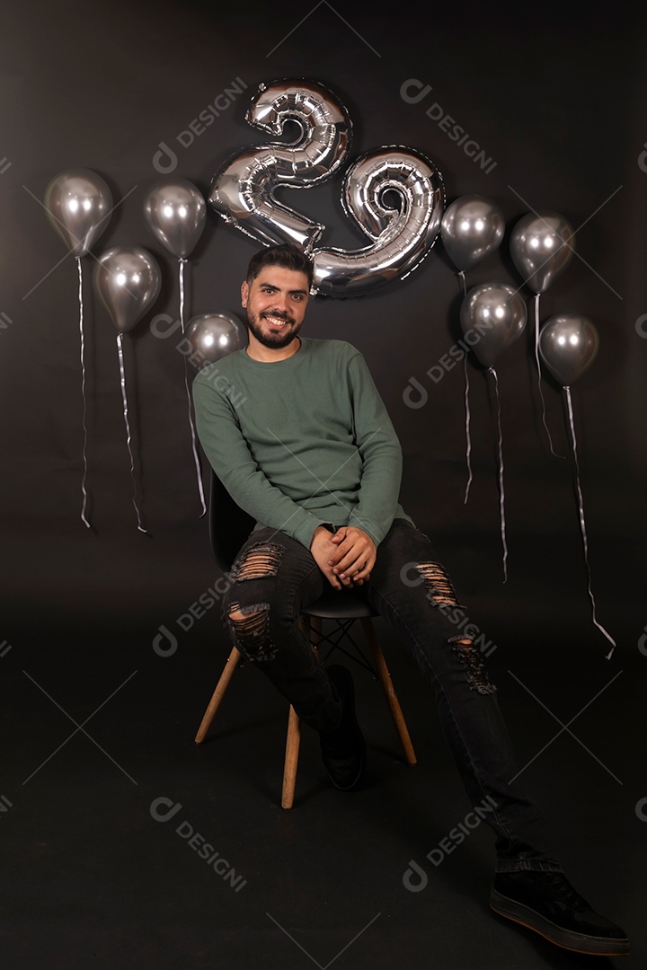 Lindo homem sentado sorridente fazendo aniversário sobre fundo com balões isolado