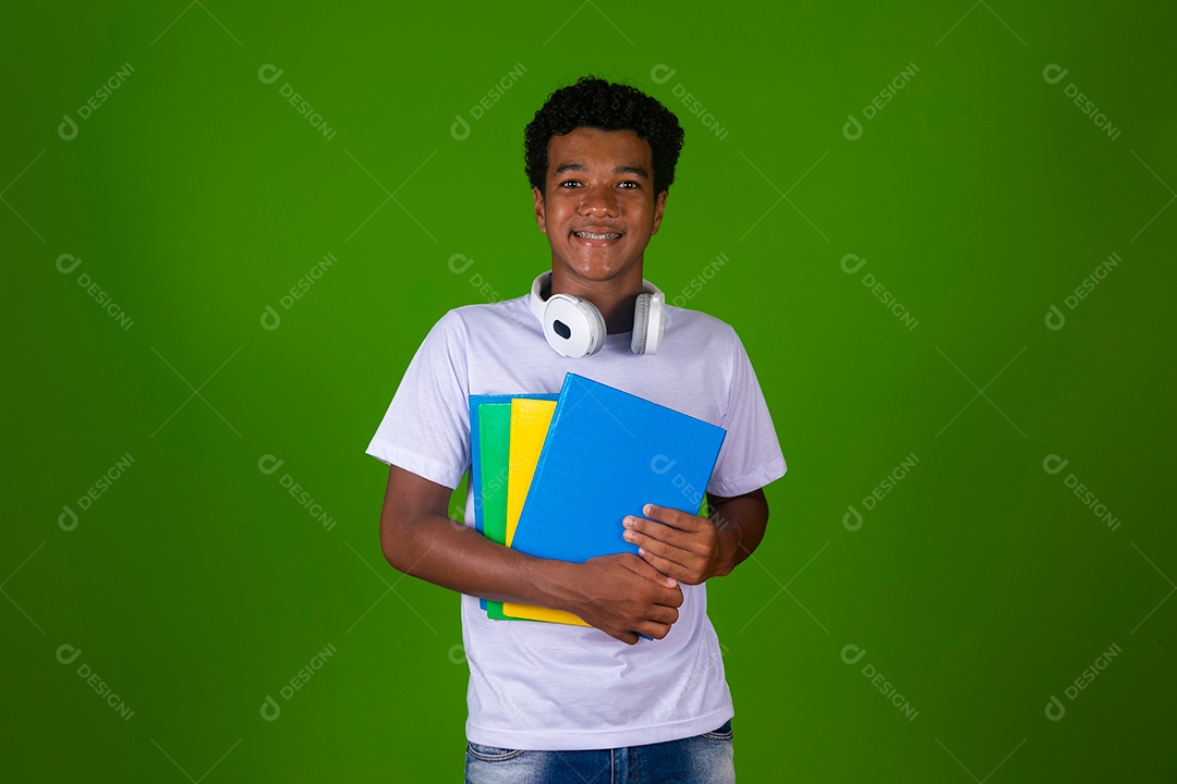 Lindo garoto negro de camisa branca usando bolsa e fone de ouvido segurando caderno escolares sobre fundo verde isolado