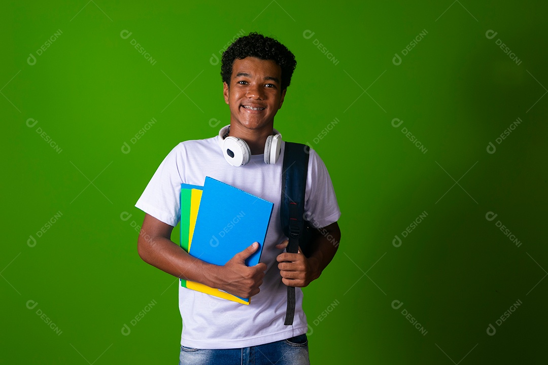 Lindo garoto negro de camisa branca usando bolsa e fone de ouvido segurando caderno escolares sobre fundo verde isolado