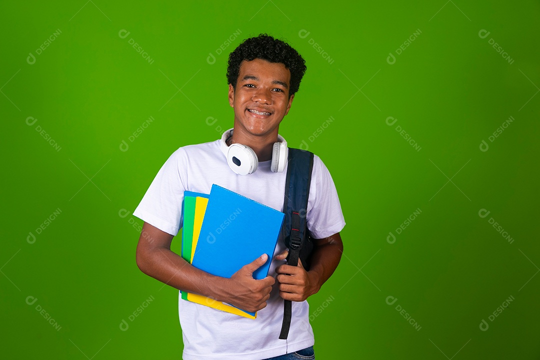 Lindo garoto negro de camisa branca usando bolsa e fone de ouvido segurando caderno escolares sobre fundo verde isolado