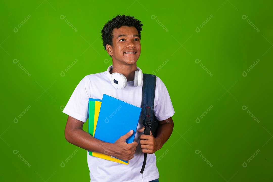 Lindo garoto negro de camisa branca usando bolsa e fone de ouvido segurando caderno escolares sobre fundo verde isolado