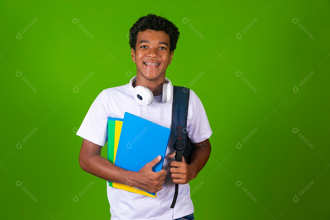 Lindo garoto negro de camisa branca usando bolsa e fone de ouvido segurando caderno escolares sobre fundo verde isolado