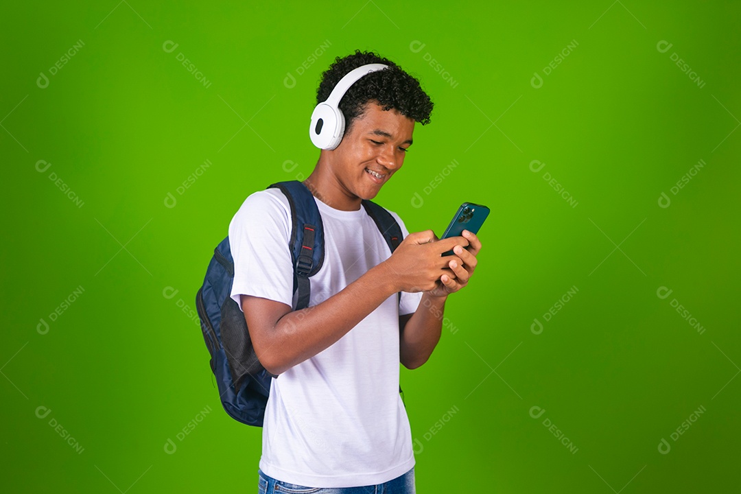 Lindo garoto negro mechendo em  smartfhone de camisa branca usando bolsa e fone de ouvido sobre fundo verde isolado
