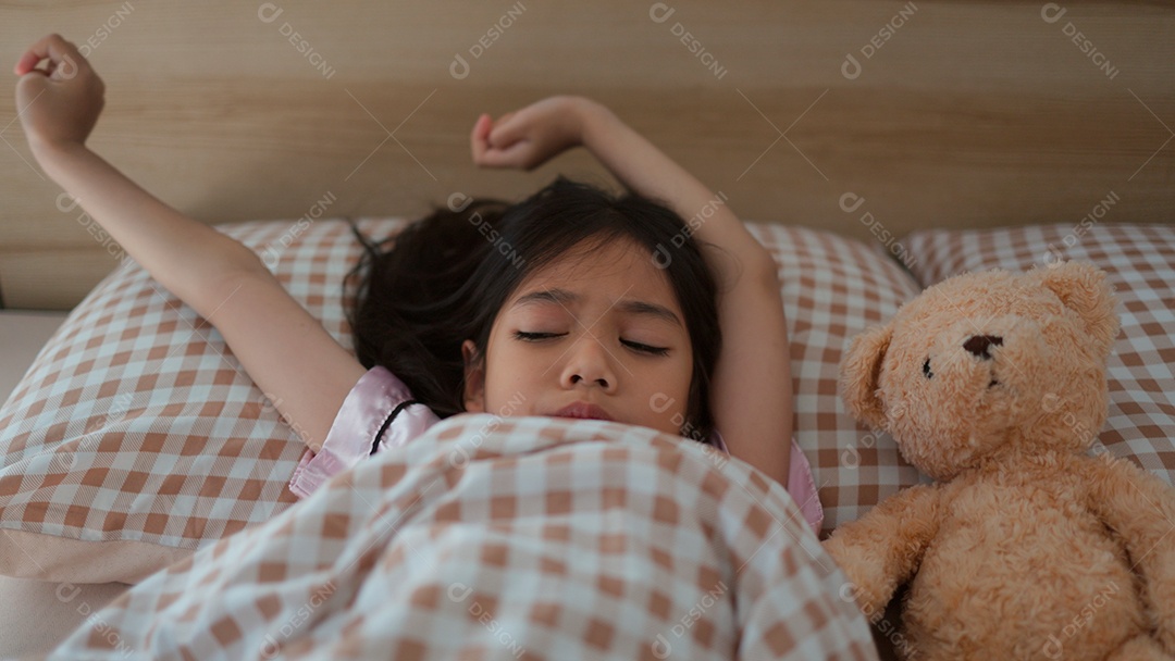 Linda menina acordando sobre sua cama ao lado do seu ursinho de pelúcia