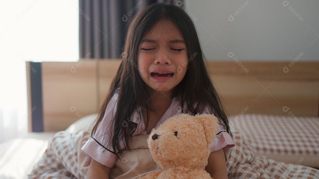 Linda menina sentada sobre sua cama assustada segurando seu ursinho de pelúcia