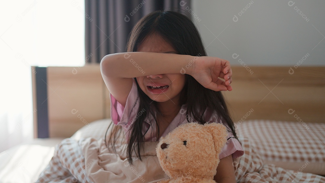 Linda menina sentada sobre sua cama assustada segurando seu ursinho de pelúcia