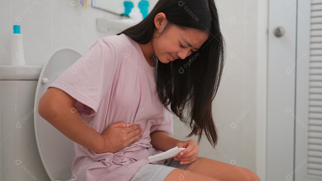 Uma menina asiática doente sentada no vaso sanitário do banheiro e segurando seu estômago doloroso durante o período menstrual