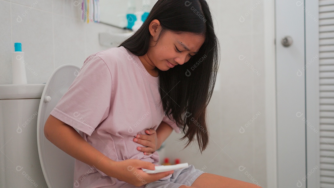 Uma menina asiática doente sentada no vaso sanitário do banheiro e segurando seu estômago doloroso durante o período menstrual